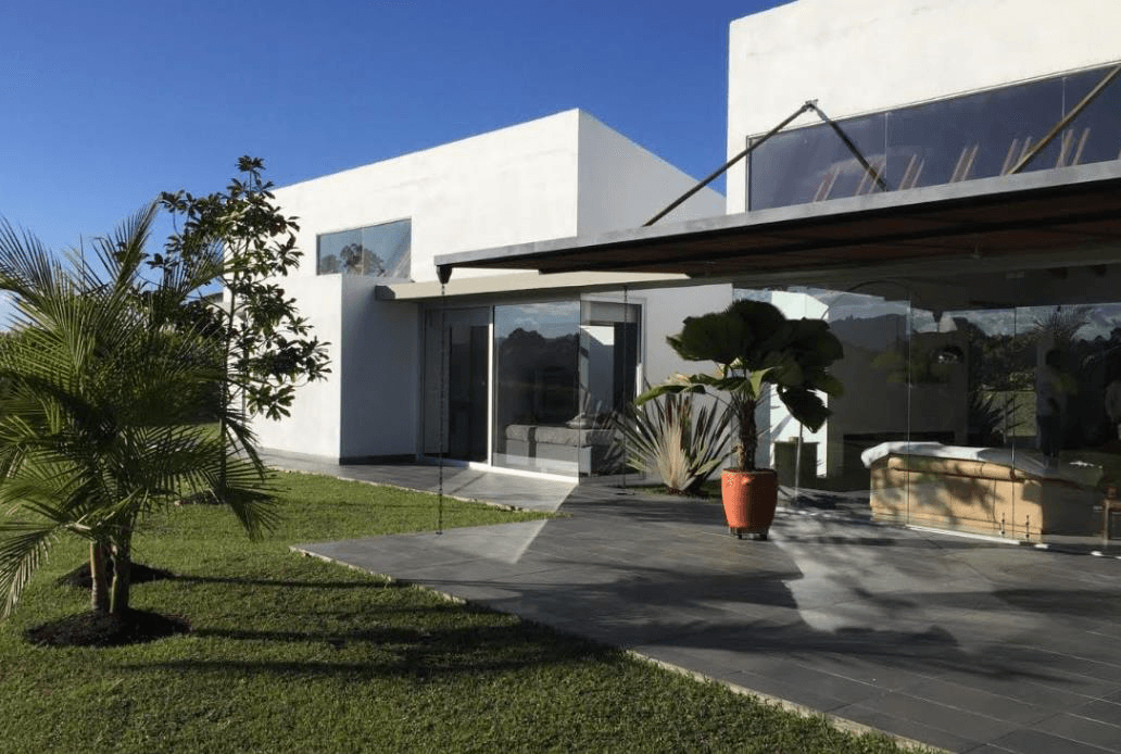 Casa San Antonio de Pereira - Fachada moderna blanca con ventanales de vidrio, terraza con baranda de cristal y jardín tropical